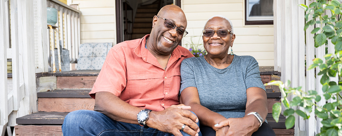 A senior couple in tinted eyeglasses, seated on their sunny front porch steps, embracing and smiling.